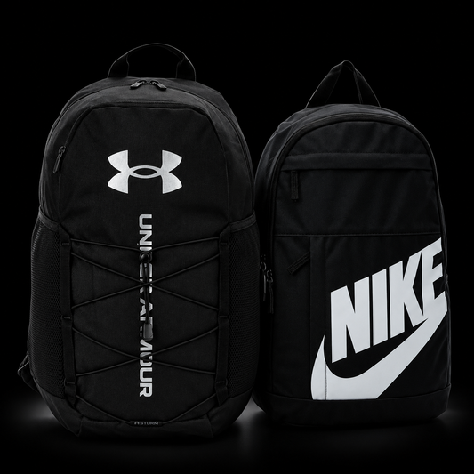 Two black backpacks shown side by side on a black background. The left backpack is Under Armour with a white logo and vertical text on the front.