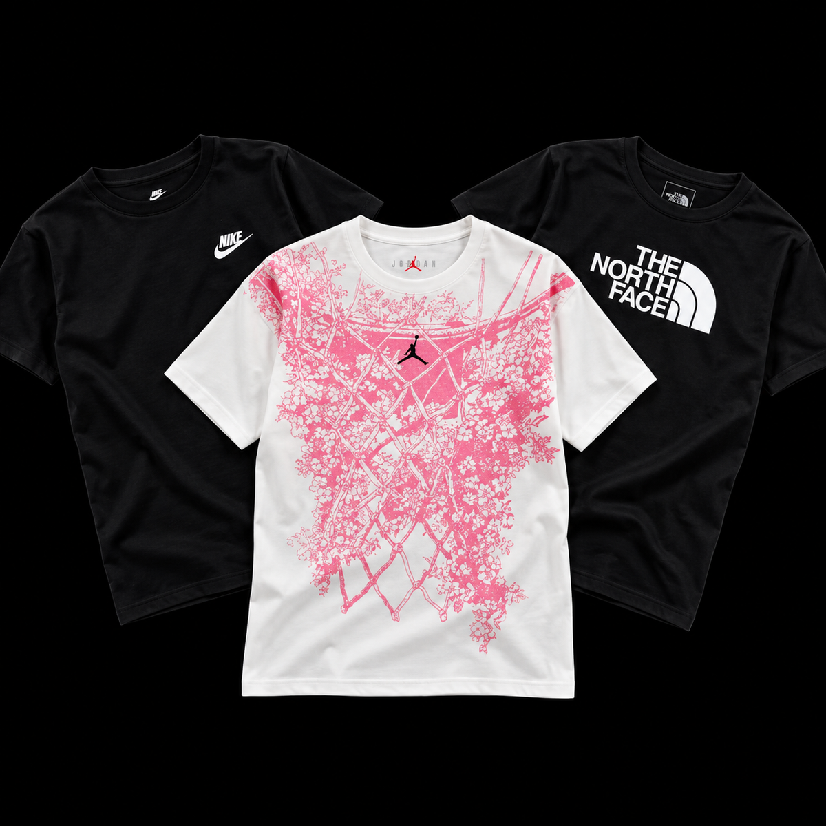 Three graphic t-shirts arranged on a black background: a black Nike t-shirt on the left, a black The North Face t-shirt on the right, and a white t-shirt in the center featuring a large pink basketball net graphic with a black Jordan logo.